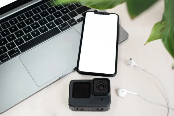 Flat lay of a laptop, smartphone with blank screen, action camera, and earphones on white background.